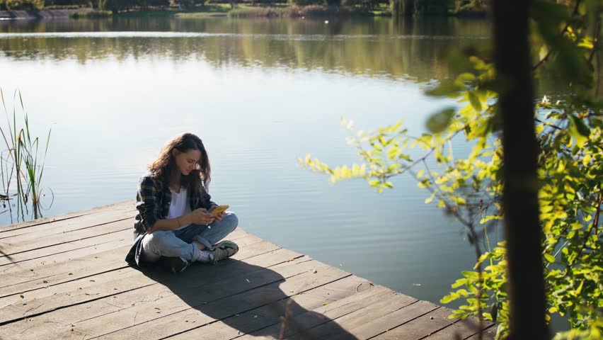 A young Caucasian woman uses her phone while sitting on a wooden pier on a lake on a sunny day.