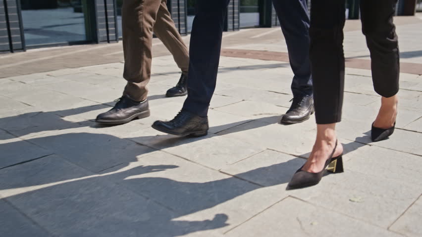 Three pairs legs walking pavement sunny day close up. Feet unrecognizable business people stepping urban street together. Unknown office colleagues strolling modern town district. Meeting concept.