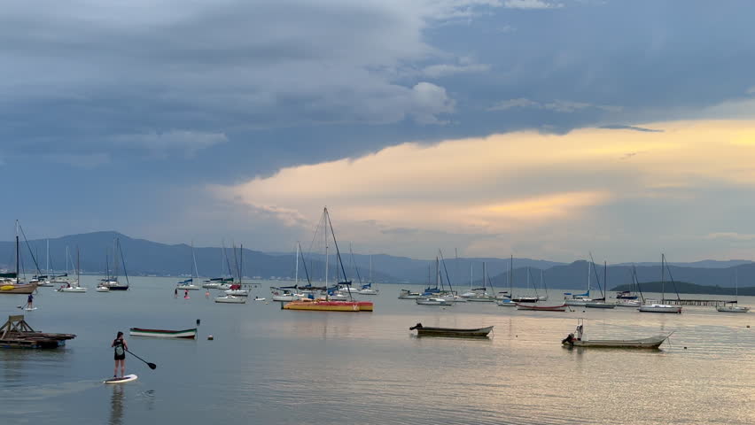 Woman on a paddle board supping on the ocean to reach a boat during a cloudy sunset. Brazil. 