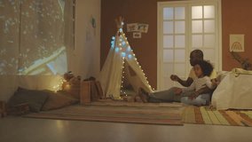Full side shot of African American father sitting on floor with son and watching educational video about bees with projector in decorated bedroom in evening