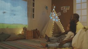 Side full footage of African American father talking about bees sitting with sleepy son on floor in decorated room with teepee while watching educational video with projector in evening