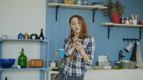 Attractive happy girl dancing and singing in kitchen while using smartphone and listening to music at home in the morning