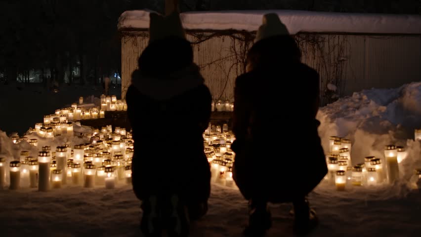 Two girls' silhouettes at the backdrop of candle lights on graves and tombstones at a cemetery. Lit candles illuminate the graves at a Christian cemetery at night. In the memory of victims of the war  Royalty-Free Stock Footage #3405804713
