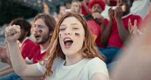 Caucasian pretty female with red hair and two German flags painted on her cheeks cheering at stadium. Video recording herself with one hand. Jumping and singing something. Showing positive emotions.