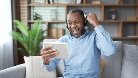 Excited mature african american male in wireless headphones watching sports match competition on tablet sitting on sofa in living room at home. Happy senior man emotionally cheering for favorite team - Powered by Shutterstock - Get 15% off with code: PIKWIZARD15