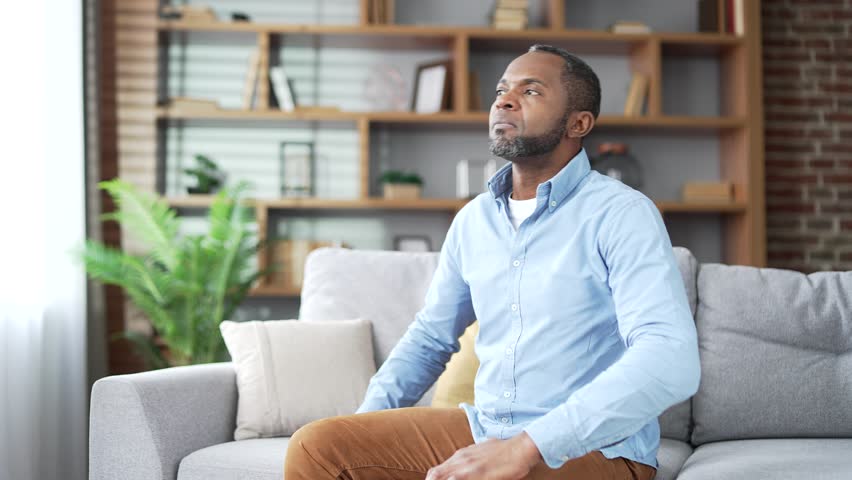 Tired mature african american male suffering from back pain while sitting on sofa in living room at home. Upset senior bearded black man holds his lower back with his hands, massages painful muscles Royalty-Free Stock Footage #3405967309