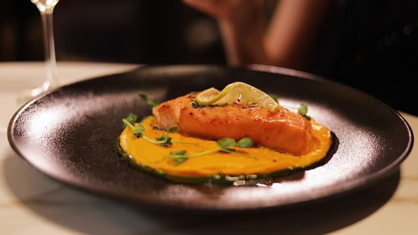 Close up of woman sitting at table at restaurant and cutting seafood dish with fork and knife in elegant way. Grilled salmon fillets served on black round plate with potato puree and pea sprouts.