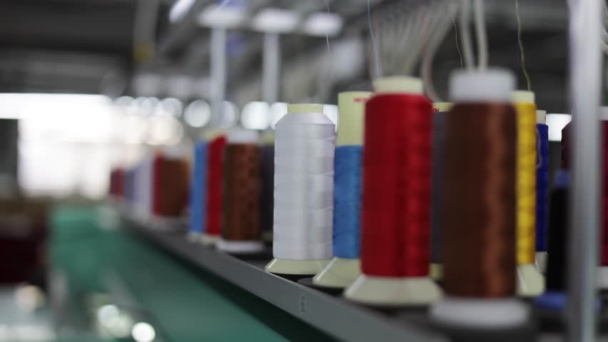 Multicolored coils of synthetic textile thread running at industrial weaving manufacturing machine. Selective focus on white sewing spool getting drawn at textile fabric. Concept of production.