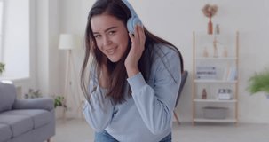 Young happy joyful smiling girl wearing casual clothes dancing in the living room listening to music in headphones. Beautiful woman having fun at home. People emotions concept. 4k video.