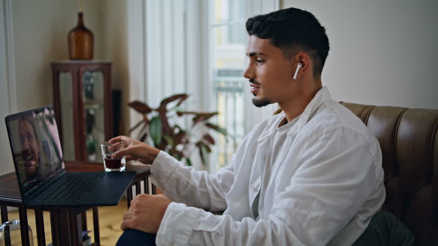 Resting guy drinking alcohol at armchair place closeup. Smiling man using earphones having laptop video call at home. Positive male person relaxing alone having virtual conversation with friend