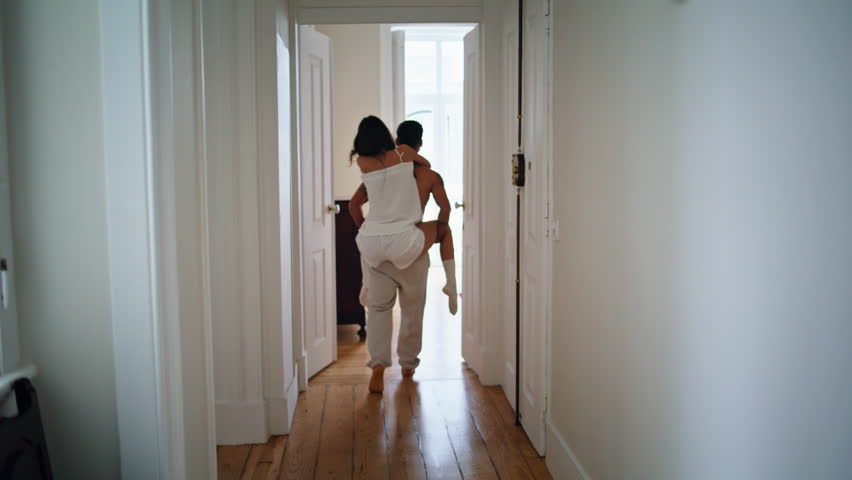 Young man piggyback wife at cozy house. Married lovers walking hall at luxury apartment. Positive newlyweds having fun at lazy weekend morning. Cute spouses relaxing together at light colours interior Royalty-Free Stock Footage #3406129407