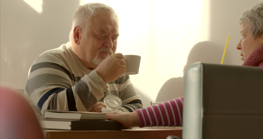 An elderly man enjoys a cup of tea or coffee in a cafe. Man surrounded by the aroma of freshly brewed coffee or the soothing fragrance of hot cup of tea. Casual conversation with woman wife or friend. Royalty-Free Stock Footage #3406152387