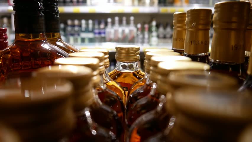 Many beautiful bottles of whiskey and brandy in alcohol store close-up and a bearded man chooses one