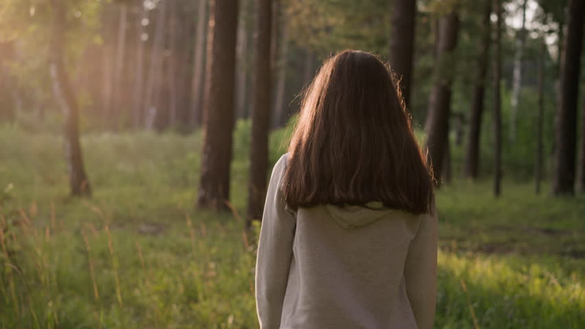 Lonely woman walking through sunny forest. Female walks alone surrounded by trees and wild environment at sunset. Lady feels empty and useless