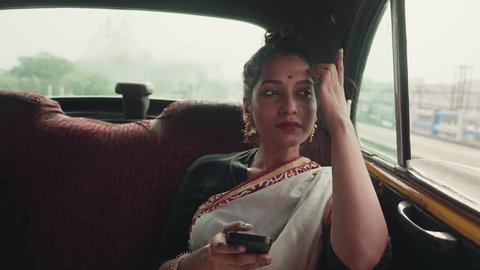 A young smiling traditional Hindu Indian Asian Girl or female in an ethnic saree is sitting in a cab and fiddling with a Mobile phone and looking around or enjoying outside view a taxi while traveling Video stock