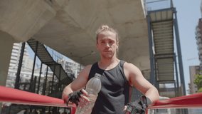 Muscular man in sportswear holding bottle of water, smiling and posing for camera at parallel bars after outdoor workout. Zoom shot, video portrait