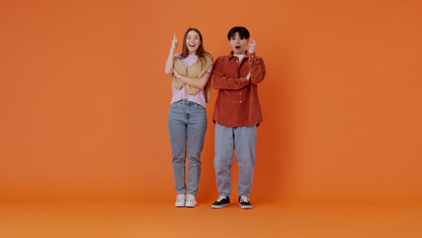 Pensive European woman and Asian man come up with idea showing Eureka finger sign. Young couple brainstorming and genial thoughts in orange room. Full size