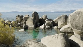 Lake beach with big stones on sunny day. Scenery view of clear river water. Lake surrounded mountains. Rocky lake shore. Huge boulders washing by river water. Natural serenity scene, 4k footage