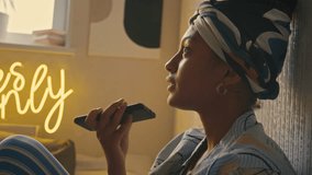 Side handheld closeup of young fashionable Black woman in hairband recording voice message on cell phone sitting at home with grunge interior in neon light