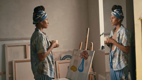 Стоковое видео: Medium handheld side footage of young Black fashionable artist in bandana drinking tea or coffee while thinking in art studio and looking out of window