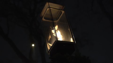 A decorative gas flame lamp illuminates above an outdoor seating area in a courtyard in the winter. Dark tree branches in the background.