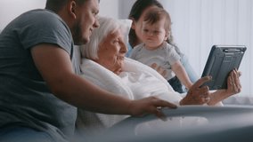 Hospital ward. Elderly woman lying in bed spends time with family. Loving relatives use digital tablet, support grandmother recovering after successful surgery. Modern medical facility. Slow motion.