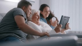 Hospital room. Old woman lying in bed spends time with relatives. Loving family use tablet computer, support elderly grandmother recovering after successful surgery. Modern medical facility or clinic.