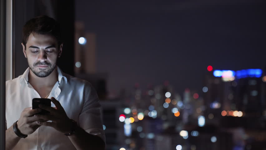 A Young Handsome Man Smiles While he Sends a Voice Message Using His Smartphone, Balcony at Night with Tall Building Skyline Lights. Man With Phone Texting and Chatting with App. Technology. Copyspace Royalty-Free Stock Footage #34073938