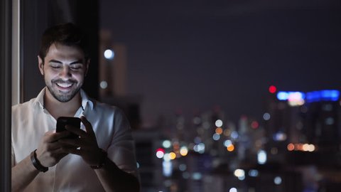 A Young Handsome Man Smiles While he Sends a Voice Message Using His Smartphone, Balcony at Night with Tall Building Skyline Lights. Man With Phone Texting and Chatting with App. Technology. Copyspace