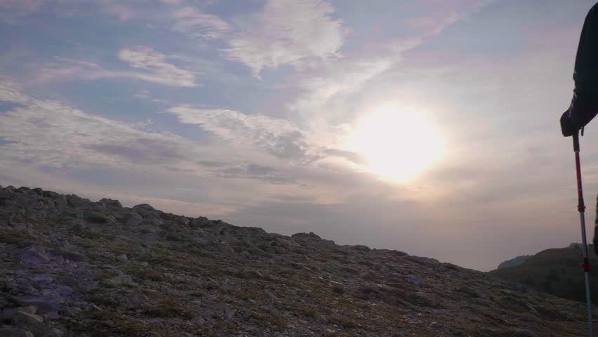 Silhouette of tired hiker man with hiking poles walking in mountains. Sun rays and blue sky. Slow motion. Steadicam low angle full shot.
