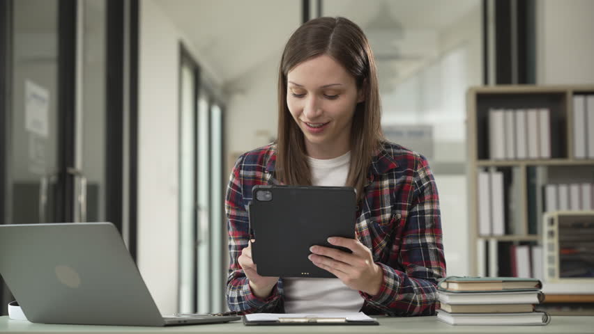 Happy young Caucasian woman university student with long brown hair, smiling while using laptop in a library setting, visiting public library for work on study research project.