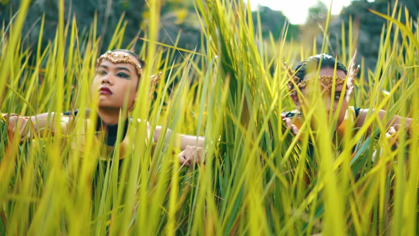 Close view through tall grass blades of two female warriors hiding low to the ground, looking around cautiously.