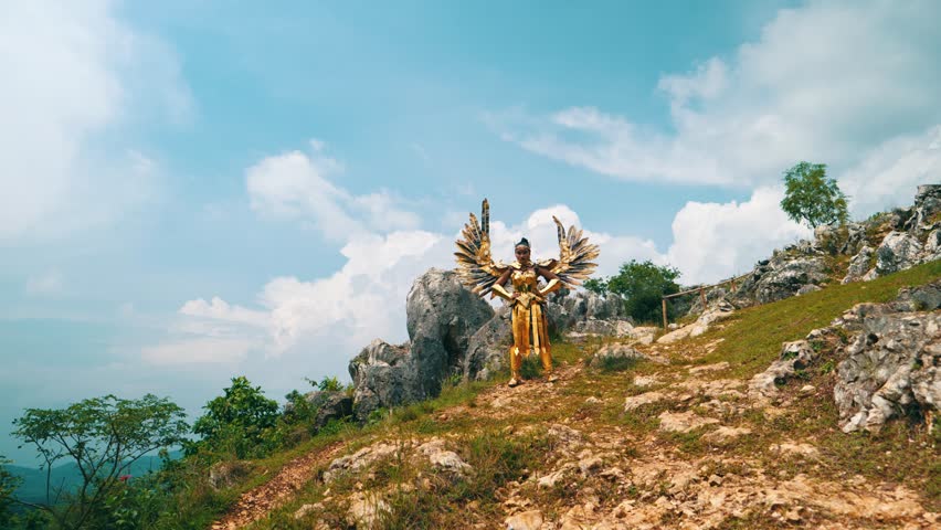 Wide shot of a majestic female warrior in full golden armor with large spread wings, posing powerfully on a rocky mountainside under a cloudy sky.