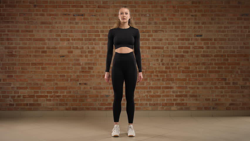 A young female trainer conducting a sports video lesson. She performs the squat side step exercise, demonstrating precisely how to do it correctly. The girl is in black sportswear. RED V-RAPTOR 8K RAW Royalty-Free Stock Footage #3407789797