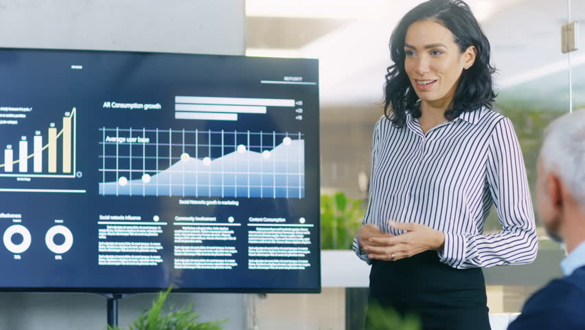 Beautiful Businesswoman Gives Report/ Presentation to Her Business Colleagues in the Conference Room, She Shows Graphics, Pie Charts and Company's Growth on the Wall TV. 