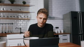 Young bored man reluctant to eat pushing a bowl full of cereal while sitting in the kitchen watching a video on a tablet computer