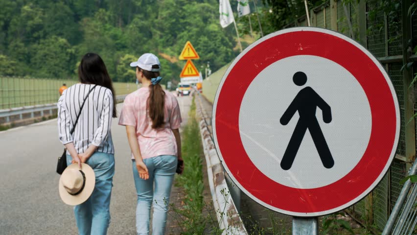 road sign, round, with black silhouette of a man in the center and a red frame, prohibiting pedestrian traffic. close-up. there are people in background. two girls walking along bridge road. repair