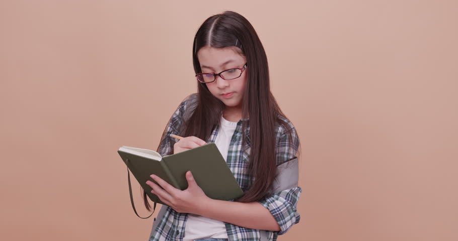Schoolgirl thinking something and write it down in a notebook. Isolated on brown background in studio.