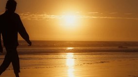 Silhouette of man walking barefoot on beach at sunset. Back view of guy enjoying sea landscape in golden hour. Male tourist looking sunrise on sandy shore. Ocean waves rolling on coast in slow motion 