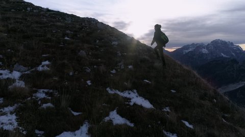 Aerial - Young woman hiking uphill on the edge of mountain in Julian Alps at sunset. Sporty girl in form fitting bottoms with a backpack hikes outside on a cold, sunny day in autumn (slow motion)