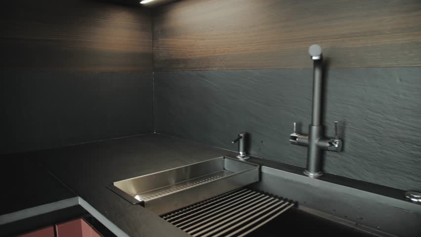 Kitchen sink and faucet, kitchen interior in dark colors. Close-up modern kitchen interior. Black ceramic tiles on the wall and countertop and wooden facade, slow motion shot.