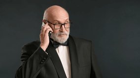 Dignified man in black tuxedo with bow tie, talking on the phone. He has a gray beard, bald head, and glasses. He stands against a dark gray background. Elderly man. RED 8K RAW. - Powered by Shutterstock - Get 15% off with code: PIKWIZARD15