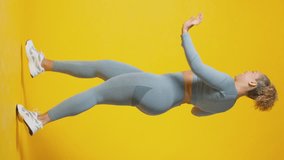 Vertical video studio full length shot of smiling woman wearing gym fitness clothing exercising on yellow background - shot in slow motion