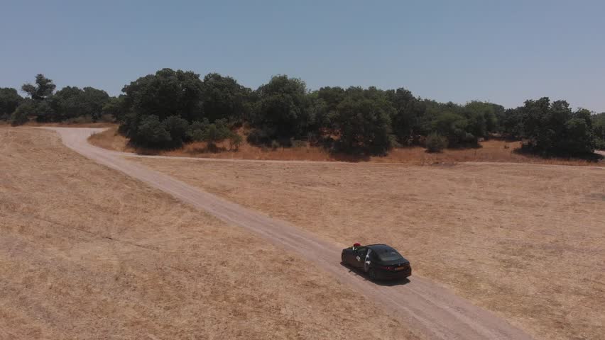 A black wedding car is driving along a dirt country road along the forest. Summer day, blue, cloudless sky.