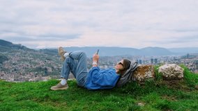A young happy smiling girl lying on the green grass on the top of hill overlooking a European city. Relaxed woman traveller using smartphone, making photo and video of her journey.