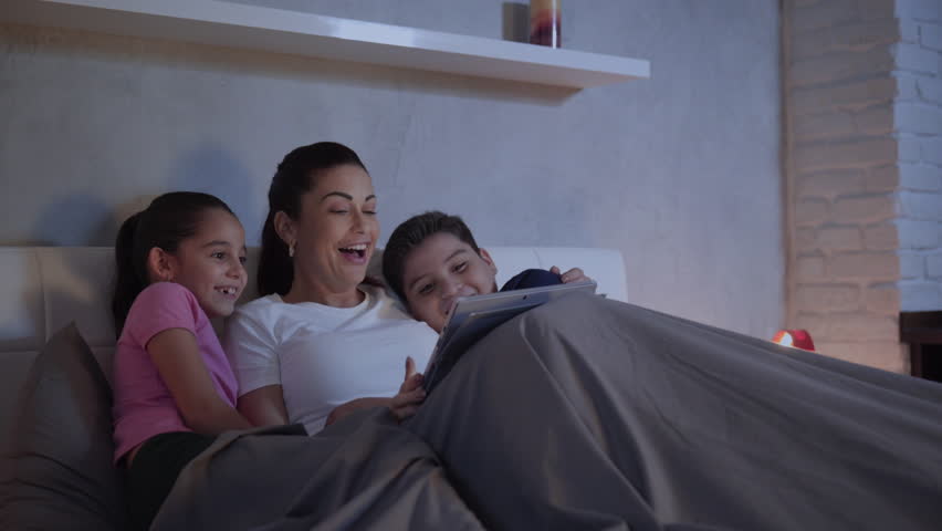 A mother and her children in bed enjoy a streaming movie in their tablet during a weekend night. A single caucasian woman and her kids having fun spending quality time together.  Royalty-Free Stock Footage #34086679