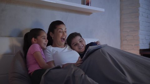 A mother and her children in bed enjoy a streaming movie in their tablet during a weekend night. A single caucasian woman and her kids having fun spending quality time together. 