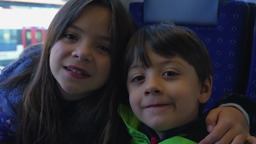 Young siblings posing for camera, 8 year old sister putting arm around her 5 year old brother while riding train looking at camera smiling. Portrait closeup faces of children Royalty-Free Stock Footage #3408675553