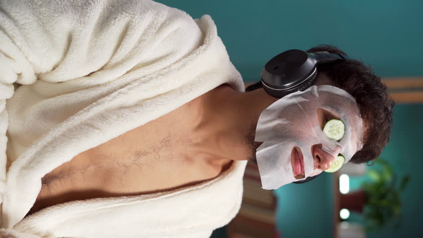 Skin care concept. Muslim man in bathrobe enjoying spa day at home and relaxing in headphones with facial beauty mask on face and fresh cucumber slices on eyes.