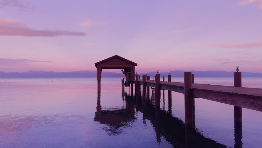 Wooden pier on the lake in sunset light. Scenic shot of old jetty at the dusk. Amazing sky in pink and violent colors. Mountains surrounded lake Water ripples on lake surface at evening. , 4k footage